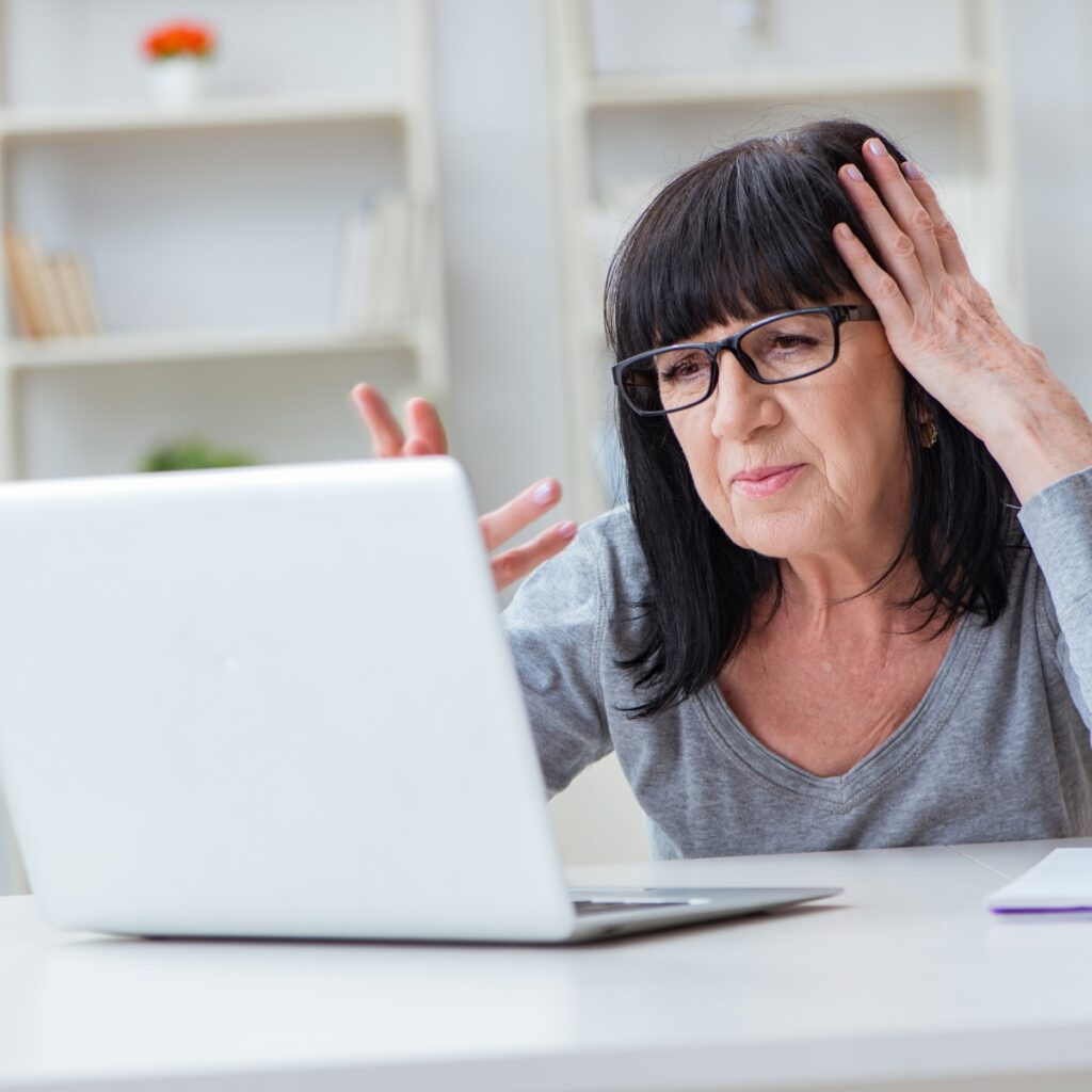 Zu sehen ist eine Frau mit schwarzen Haaren und einem grauen T Shirt. Sie sitzt vor dem Notebook und ist gestresst.